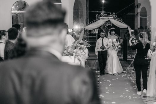 Father walks smiling bride down the aisle at indoor wedding ceremony. Captured in black and white.