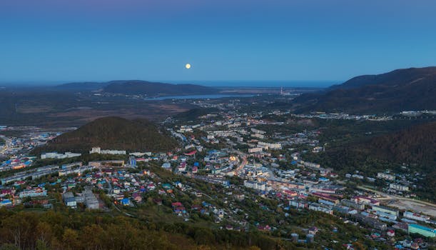 Aerial cityscape featuring mountains, a full moon, and vibrant rooftops under a clear evening sky.
