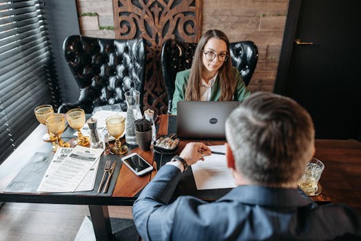 A professional business meeting indoors with two adults discussing and working on laptops.