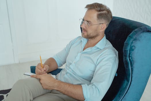 A therapist in a session, wearing glasses and taking notes while seated on an armchair.
