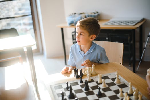 Boy in a blue shirt intently playing chess at a wooden table in a bright room.