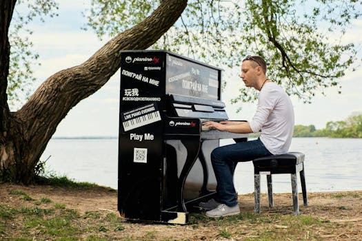 A man sits playing an outdoor piano near a tranquil lakeside, surrounded by trees.