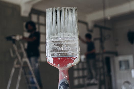 Detailed close-up of a paintbrush with men painting a wall in the blurred background, highlighting renovation work.