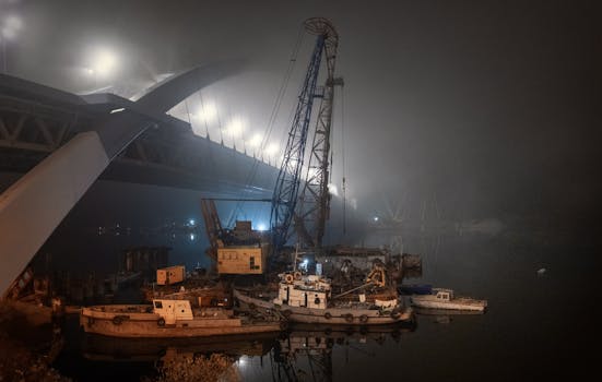 Misty night at a shipyard with a towering crane and illuminated bridge.