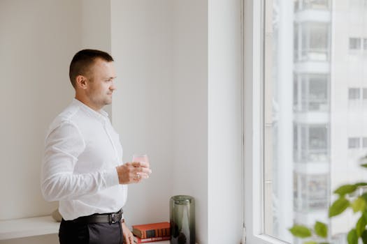 Adult man in white shirt holding a glass, looking through the window indoors.