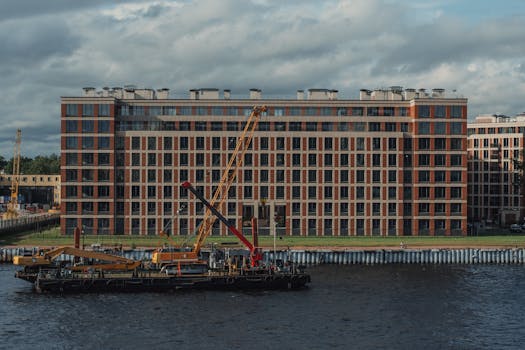 Contemporary multistory masonry house facade against city river with hoisting crane on pontoon under cloudy sky